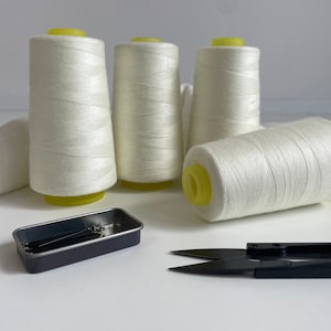 Könnte beinhalten: Vier weiße Garnrollen mit gelben Oberteilen und ein kleiner schwarzer Metallbehälter mit Nähnadeln.