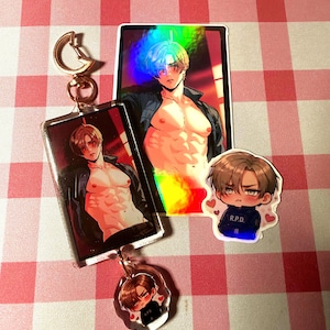 Puede incluir: Un llavero rectangular de acrílico con un personaje de anime sin camisa y cabello castaño, unido a un broche dorado en forma de luna. Se incluyen una pegatina holográfica rectangular y una pequeña pegatina circular de un personaje chibi con una camiseta R.P.D.