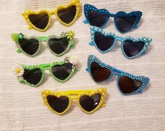 May include: Six pairs of heart-shaped sunglasses with jeweled embellishments. The sunglasses are in various colors, including yellow, green, and blue.