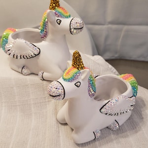 May include: Two white unicorn planters with rainbow manes, gold horns, and crystal accents. The planters have open containers for plants or storage. The unicorns have wings and are resting on a white surface.