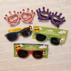 May include: Four pairs of children's sunglasses with jeweled crowns. The sunglasses are blue, green, red, and purple. The packaging includes the text "Kid's Sunglasses" and "Lunettes de Soleil pour Enfant".
