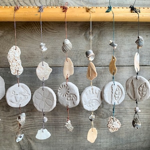 May include: Handmade wind chimes, each with a white ceramic disc featuring embossed designs like an anchor and flower. The discs are strung with shells and stones, creating a rustic, coastal aesthetic. The chimes hang from a wooden rod.