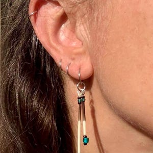 Puede incluir: Un pendiente colgante con dos púas finas y cónicas de color crema con puntas de color marrón oscuro, acentuado por pequeñas cuentas de color turquesa vibrante y negro. El pendiente cuelga de un aro de metal plateado en el lóbulo.