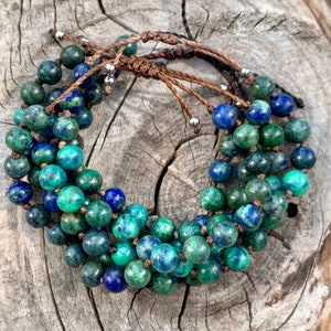 Peut inclure: Un bracelet de perles rondes et polies dans des tons de bleu et de vert. Les perles sont enfilées sur un cordon marron et fixées par un nœud coulissant. Le bracelet est présenté sur une surface en bois rustique. Les perles mesurent environ 1 cm de diamètre.