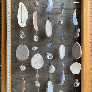 May include: A decorative wall hanging featuring various seashells and ceramic pieces strung on twine. The arrangement includes white, beige, and blue shells, along with carved ceramic pendants. The wooden frame measures approximately 50 cm in height.