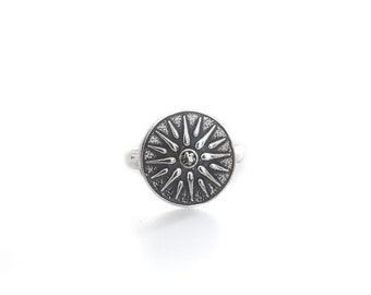 May include: A silver ring featuring a circular face with a raised sunburst design. The center of the sunburst has a small, circular detail, and the background of the face has a dark, oxidized finish to highlight the silver rays radiating outward.