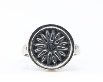 May include: A silver ring featuring a circular face with a raised sunburst design. The center of the ring has a dark, oxidized finish that highlights the metallic rays radiating outward from a small central point within the circular border.