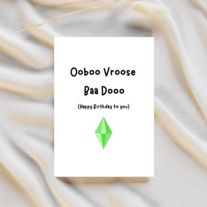 以下が含まれることがあります： 白いカードに黒い文字で「Ooboo Vroose BAA Dooo（お誕生日おめでとう）」と書かれています。テキストの下には緑色のダイヤモンドの形があります。