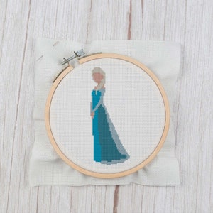 Könnte beinhalten: Ein Kreuzstich-Stickrahmen mit einem fertigen Design einer Comicfigur, die ein blaues Kleid und einen weißen Umhang trägt. Die Figur hat blonde Haare und ein hellblaues Gesicht.