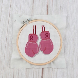 May include: Pink boxing gloves with pink ribbons on each glove, embroidered on white fabric in a wooden embroidery hoop.