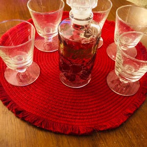 May include: A clear glass decanter with a decorative stopper, surrounded by six small stemmed glasses. The decanter and glasses sit on a round red mat. The decanter appears to contain a red liquid.