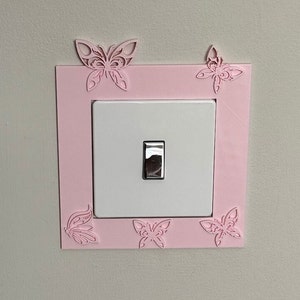 May include: A pink light switch cover with a white light switch and butterfly cutouts around the edges.