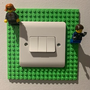 May include: A white light switch with two buttons is surrounded by a green Lego-style baseplate. Two Lego minifigures are attached to the baseplate.