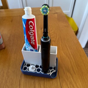 May include: A white and blue toothbrush holder with a blue base. A tube of Colgate toothpaste and an electric toothbrush are in the holder.