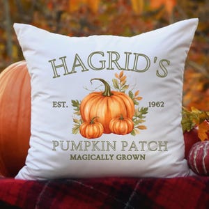 May include: White square pillow with the text "Hagrid's Pumpkin Patch" and a watercolor illustration of pumpkins and leaves. The pillow has the text "Est. 1962" and "Magically Grown". The background is blurred with fall colors.