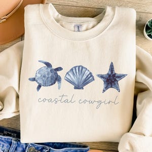 May include: A cream-colored sweatshirt with a blue watercolor design of a sea turtle, seashell, and starfish. The text "coastal cowgirl" is printed below the design.