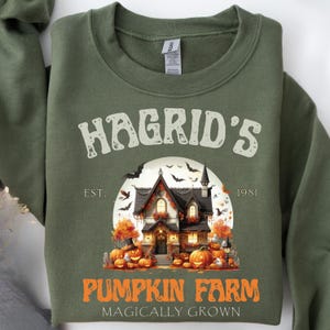 Może przedstawiać: Zielona bluza z nadrukiem domu, dyni i nietoperzy oraz napisem "Hagrid's Pumpkin Farm" w kolorze pomarańczowym i białym. Projekt zawiera tekst "EST. 1981" i "Magically Grown".