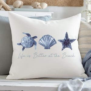May include: White square pillow with a blue watercolor design of a sea turtle, seashell, and starfish. The text "Life is Better at the Beach" is printed below the illustrations. The pillow is a decorative item.