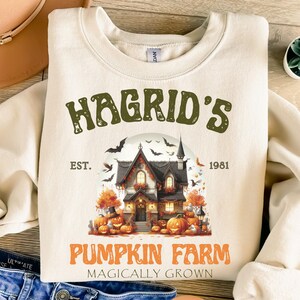 May include: Cream-colored sweatshirt featuring the text "Hagrid's Pumpkin Farm" with a graphic of a spooky house, pumpkins, and bats. The text "Est. 1981" and "Magically Grown" are also included. A festive Halloween-themed design.