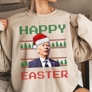 Könnte beinhalten: Beiges Sweatshirt mit einer Grafik von Joe Biden mit Weihnachtsmütze. Die Worte "HAPPY" und "EASTER" sind in grünem und rotem Pixeltext mit Weihnachtsbaum- und Punktmustern dargestellt.