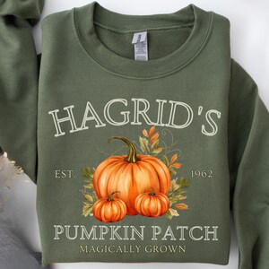Könnte beinhalten: Olivgrünes Sweatshirt mit dem Text "Hagrid's Pumpkin Patch" und einer Illustration von Kürbissen und Herbstlaub. Das Design enthält den Text "EST. 1962" und "Magically Grown".