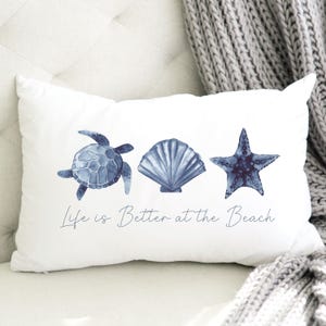 May include: White rectangular pillow featuring watercolor illustrations of a blue sea turtle, seashell, and starfish. The text "Life is Better at the Beach" is written below the illustrations. The pillow is on a white surface.