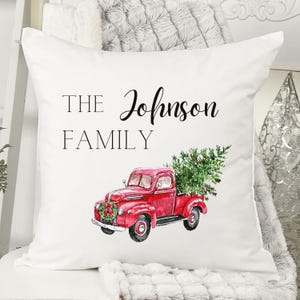 May include: White cushion with the text "THE Johnson FAMILY". It features a watercolour illustration of a red vintage truck carrying a Christmas tree. A festive wreath adorns the front of the truck.