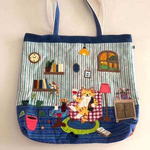 Könnte beinhalten: Eine blau-weiß gestreifte Tragetasche mit einem skurrilen Katzendesign. Die Katze liegt in einem Stuhl und hält ein Weinglas und eine Flasche. Die Tasche hat Applikationen, darunter eine Uhr, Bücher und ein Fenster.