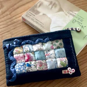 May include: A navy blue quilted pouch with a patchwork design of floral and patterned fabrics. A blue ribbon and decorative stitching adorn the edges. The pouch has a small label with the initials "G.R." and three pink buttons.