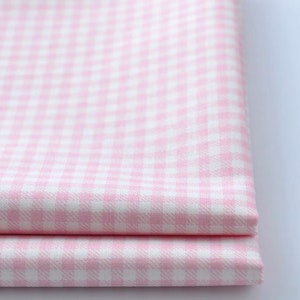 May include: Close-up of two folded pieces of pink and white gingham fabric. The fabric has a classic checkered pattern, with small squares of pink and white. The fabric appears to be cotton or a similar material.
