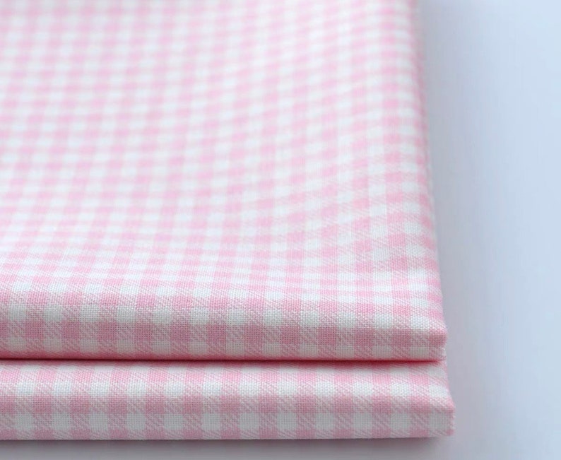 May include: Close-up of two folded pieces of pink and white gingham fabric. The fabric has a classic checkered pattern, with small squares of pink and white. The fabric appears to be cotton or a similar material.