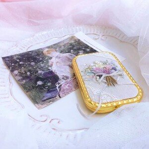 Puede incluir: Un peque&ntilde;o estuche rectangular dorado con un interior de tela blanca que presenta un dise&ntilde;o de bordado floral. El estuche est&aacute; sobre una superficie blanca con una superposici&oacute;n de encaje blanco.