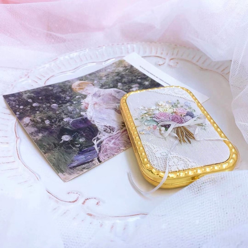 Puede incluir: Un peque&ntilde;o estuche rectangular dorado con un interior de tela blanca que presenta un dise&ntilde;o de bordado floral. El estuche est&aacute; sobre una superficie blanca con una superposici&oacute;n de encaje blanco.