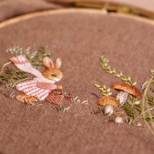 May include: Embroidered hoop art featuring a brown rabbit wearing a pink dress and white scarf, holding a basket of berries. The rabbit is surrounded by green foliage and brown mushrooms.