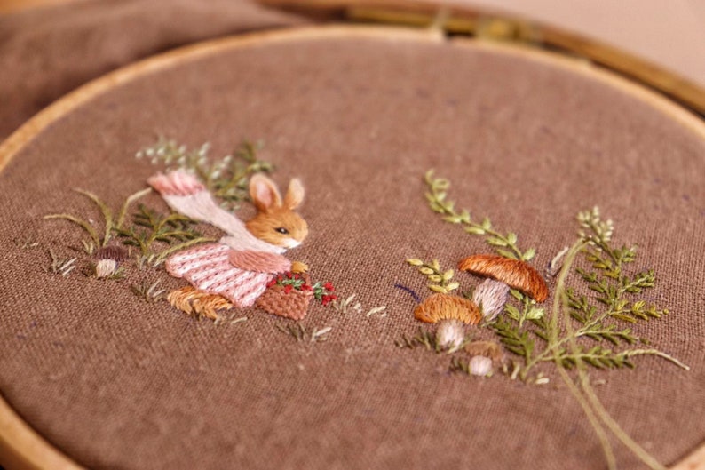 May include: Embroidered hoop art featuring a brown rabbit wearing a pink dress and white scarf, holding a basket of berries. The rabbit is surrounded by green foliage and brown mushrooms.