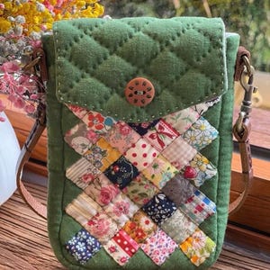 Könnte beinhalten: Eine grüne, gesteppte Umhängetasche aus Stoff mit einem Patchwork-Design auf der Vorderseite. Der Patchwork besteht aus kleinen, bunten Stoffquadraten mit Blumen- und Mustermotiven. Die Tasche hat einen braunen Knopfverschluss und einen braunen Lederriemen.