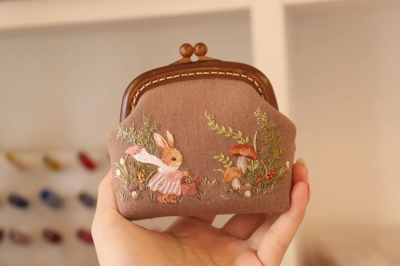 May include: A brown fabric coin purse with a wooden frame and a clasp. The purse is embroidered with a rabbit carrying a basket and mushrooms.