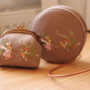 May include: Two round fabric purses with embroidered images of rabbits and mushrooms. The smaller purse has a clasp closure and the larger purse has a brown leather strap.