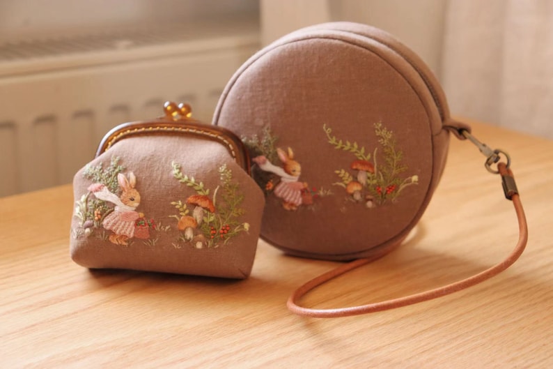 May include: Two round fabric purses with embroidered images of rabbits and mushrooms. The smaller purse has a clasp closure and the larger purse has a brown leather strap.