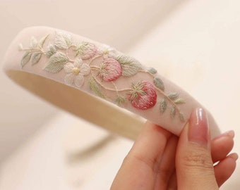 Puede incluir: Una diadema blanca delicada con fresas rosas y hojas verdes bordadas. La diadema está hecha de un tejido suave y es perfecta para añadir un toque de fantasía a cualquier atuendo.