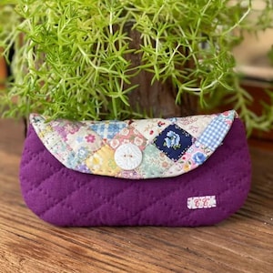 May include: A quilted purple clutch bag with a patchwork flap. The flap features floral and geometric patterns in pastel colours. A large white button secures the flap. The initials "C.C." are embroidered on the lower right corner.
