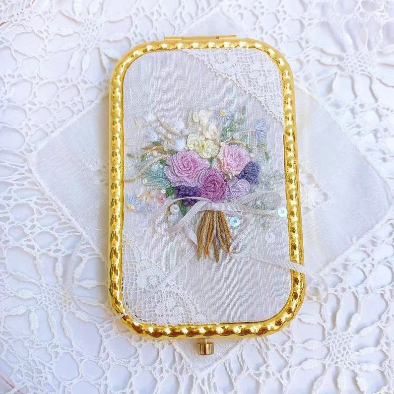 Puede incluir: Un espejo compacto dorado con un interior de tela blanca que presenta un ramo de flores bordado en tonos rosa, morado y blanco.
