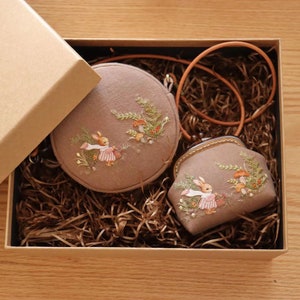 May include: Two small, round, brown purses with embroidered images of rabbits and mushrooms. The purses are in a brown box with shredded paper.