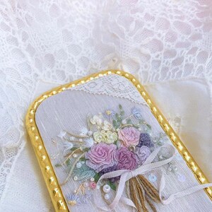 Puede incluir: Un delicado ramo bordado de flores rosas, blancas y moradas con detalles de perlas, enmarcado en un borde dorado.