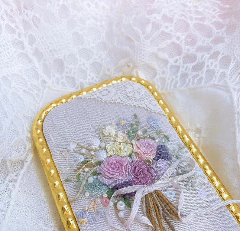 Puede incluir: Un delicado ramo bordado de flores rosas, blancas y moradas con detalles de perlas, enmarcado en un borde dorado.