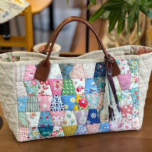 Puede incluir: Un bolso tote acolchado con un exterior de patchwork de varios patrones de tela florales y geométricos en rosa, azul, amarillo y verde. El bolso tiene paneles laterales acolchados en beige, dos asas de cuero marrón y un adorno de borla de tela.