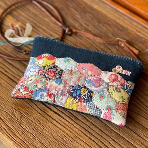 May include: A small, rectangular patchwork pouch with a navy blue top and a brown leather strap. The patchwork is made of various floral fabrics in shades of pink, blue, yellow, and red. The pouch is likely handmade and has a vintage or bohemian aesthetic.