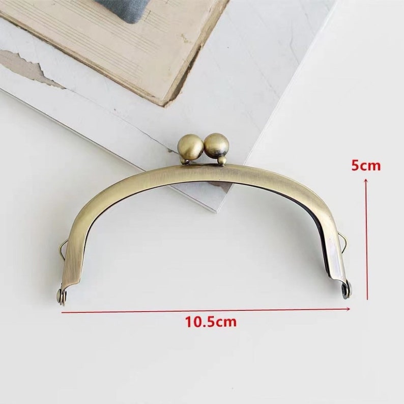 May include: A gold-toned metal purse frame with two round decorative knobs at the top. The frame measures 10.5 inches wide and 5 inches tall.
