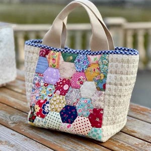 Puede incluir: Un bolso tote con un panel central de parches de tela hexagonales multicolores en varios estampados florales y geométricos. Los laterales son de tela acolchada color crema y el borde superior está rematado con un ribete de cuadros vichy azul y blanco.