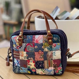 Op de afbeelding: Een marineblauwe handtas met een patchwork-ontwerp. Het patchwork heeft verschillende bloemen- en patronenstoffen in tinten rood, blauw en groen. Het heeft bruine leren handvatten en een schouderriem.